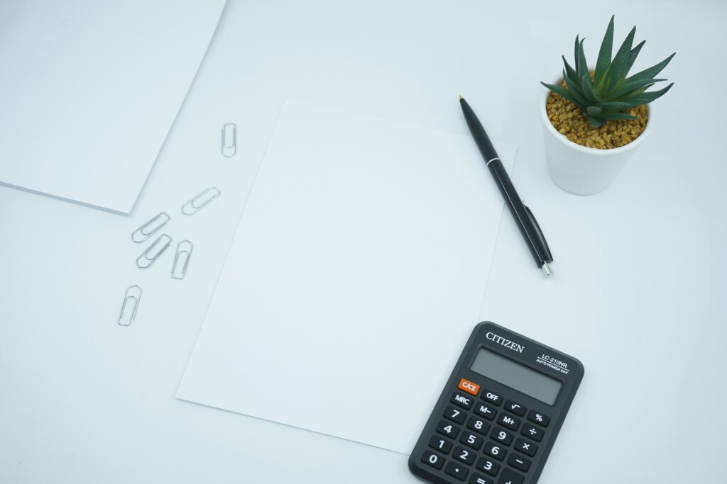flatlay of plain white paper, pen, calculator, paper pins and small plant on a white desk