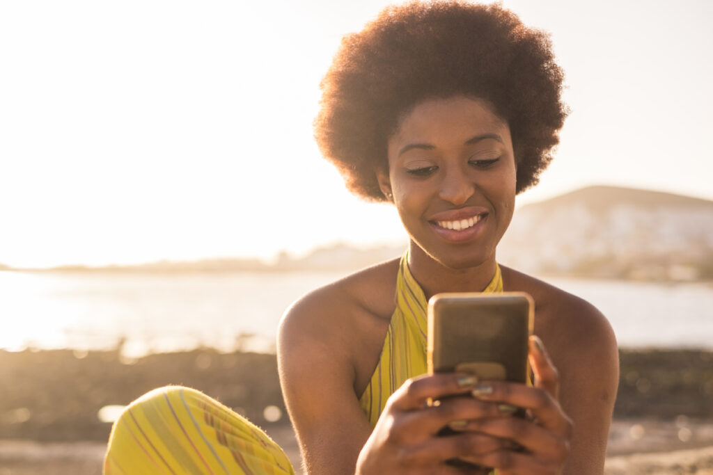 daily summer sunnny scene with black race afro american beautiful girl with alternative hair looking at the phone and check social media. sunny golden light beach and ocean in the background. vacation concept